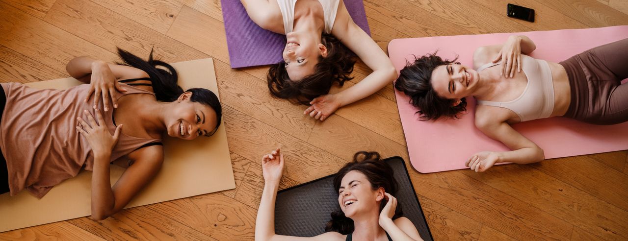 Vier Frauen liegen jeweils auf einer Yogamatte auf einem Holzboden und genießen lächelnd den erholsamen Moment nach ihrem Workout