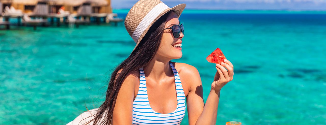 Eine Frau isst ein Stück Melone auf einem Steg und hinter ihr ist das blaue Meer