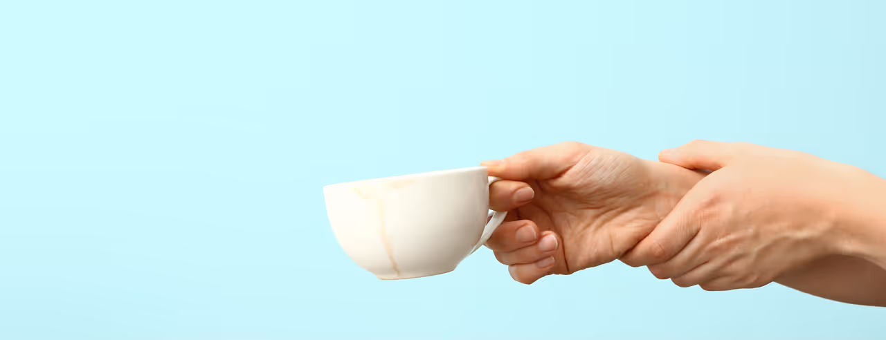 Persona con síndrome de Parkinson sostiene las manos temblorosas, bajo la taza que sostiene hay un líquido de té.