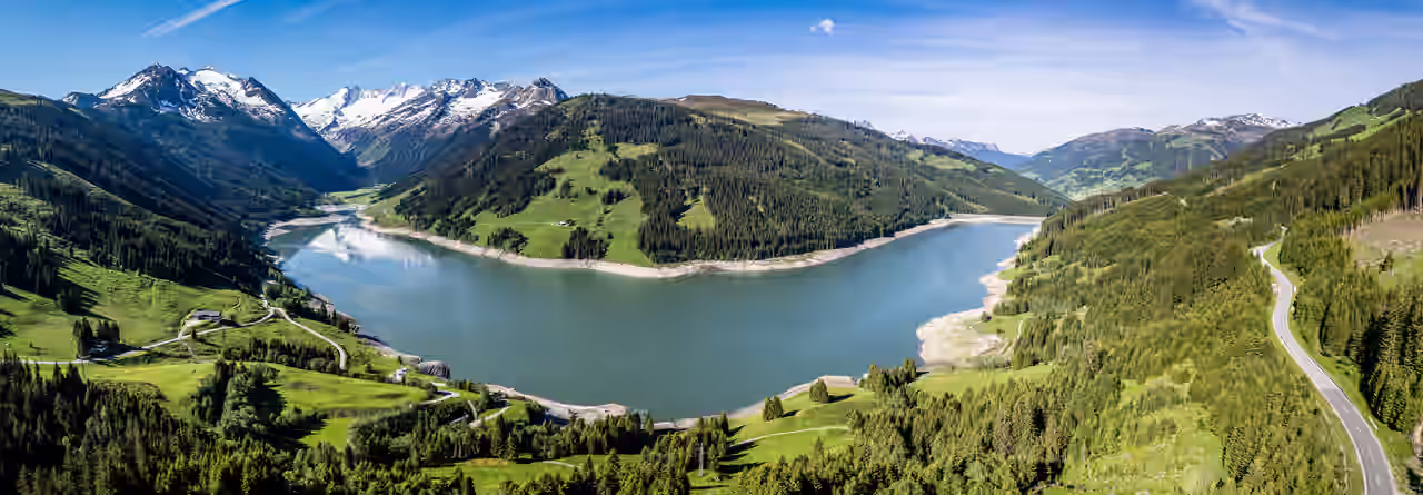 Der Durlassboden Speicher zwischen bewaldeten Berghängen, im Hintergrund die Alpen.