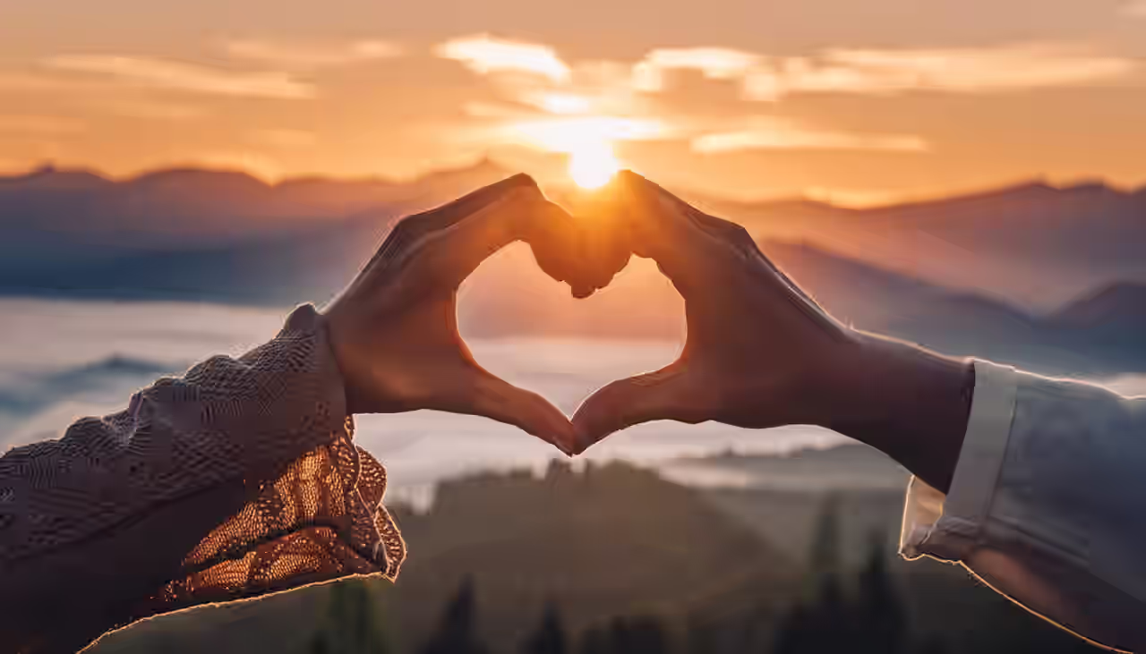 Ein Paar formt ein Herz mit den Händen. Im Hintergrund ist der Sonnenuntergang über den Bergen zu sehen.