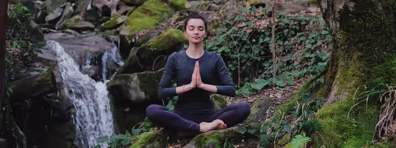 Eine Frau praktiziert Yoga inmitten eines grünen Waldes, der von Sonnenlicht geflutet ist. Die Morgensonne leuchtet zwischen all dem grün sanft in den Wald.