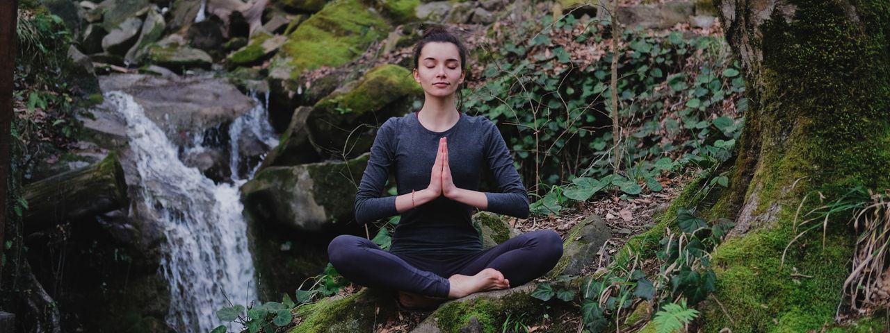 Eine Frau praktiziert Yoga inmitten eines grünen Waldes, der von Sonnenlicht geflutet ist. Die Morgensonne leuchtet zwischen all dem grün sanft in den Wald.