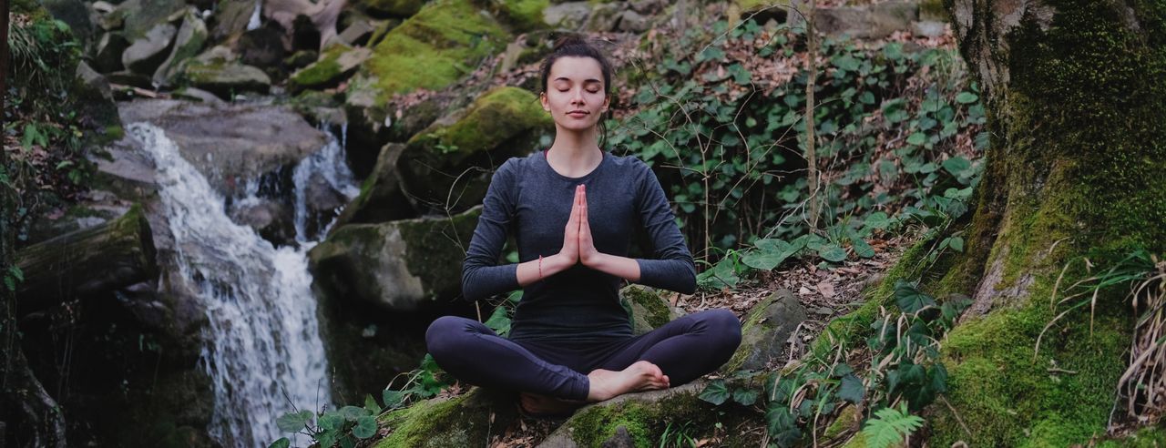 Eine Frau praktiziert Yoga inmitten eines grünen Waldes, der von Sonnenlicht geflutet ist. Die Morgensonne leuchtet zwischen all dem grün sanft in den Wald.