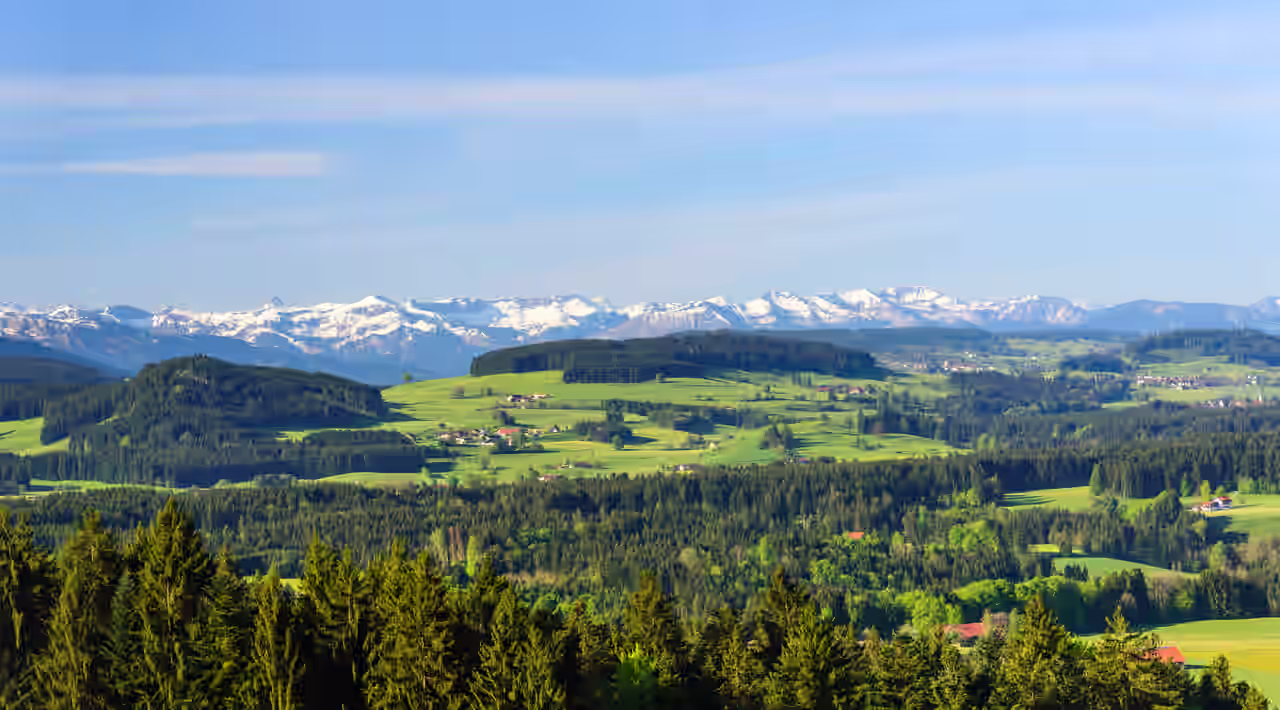 Naturlandschaft mit vereinzelten Dörfern zwischen Wiesen und Wäldern vor den schneebedeckten deutschen Alpen