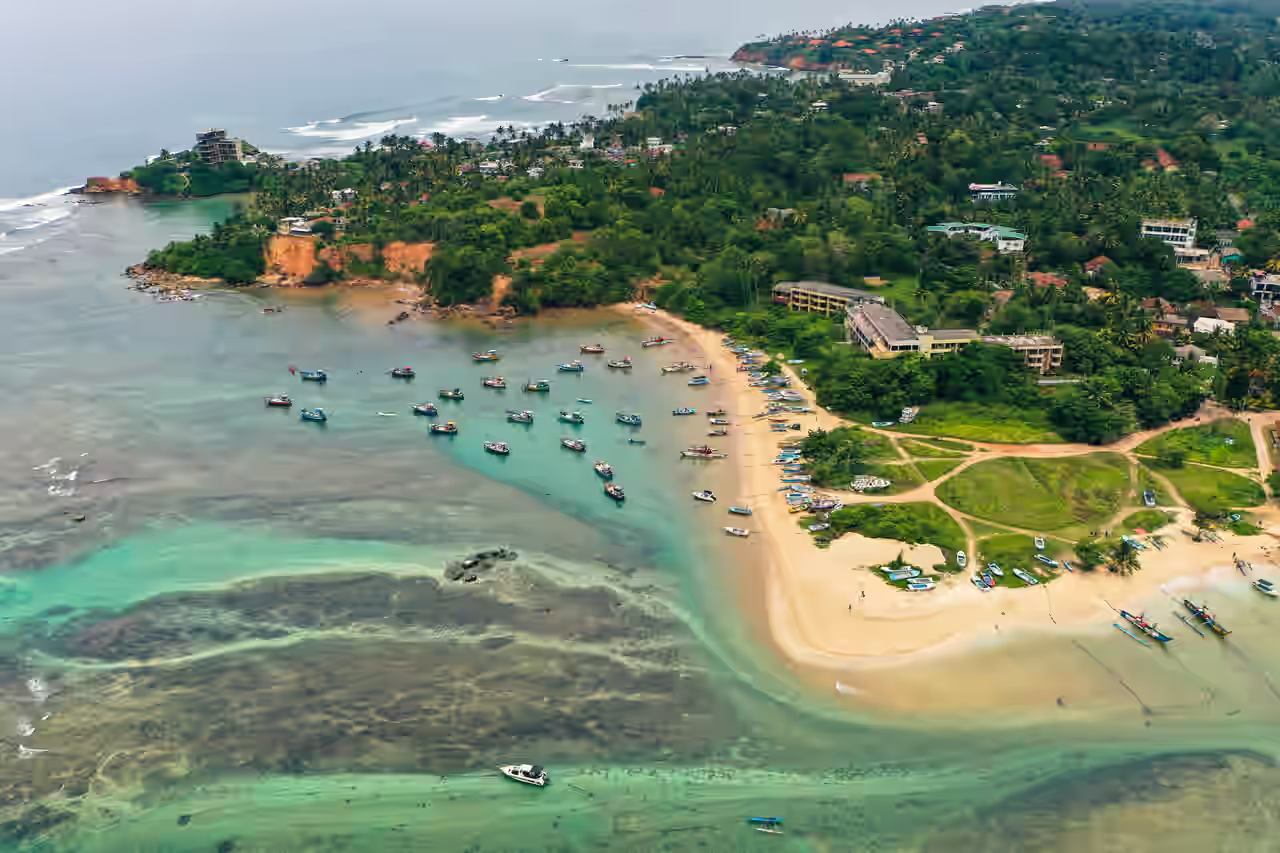 Fischerboote im Wasser am goldenen Sandstrand vor den dichten grünen Bäumen rund um Gebäude in Weligama