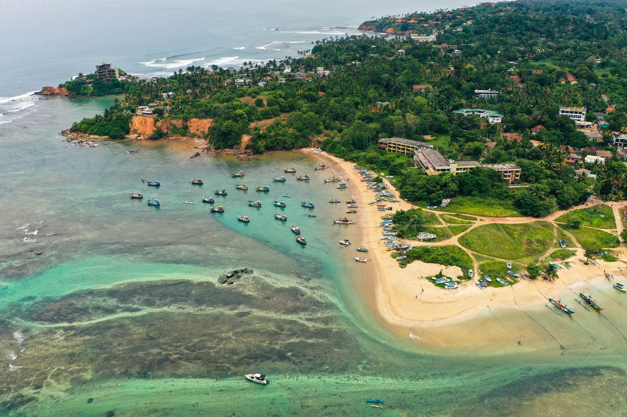 Fischerboote im Wasser am goldenen Sandstrand vor den dichten grünen Bäumen rund um Gebäude in Weligama