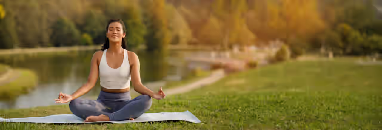 Eine Frau sitzt im Schneidersitz mit geschlossenen Augen und einem leichten Lächeln im Gesicht auf einer Yogamatte auf einer grünen Wiese.
