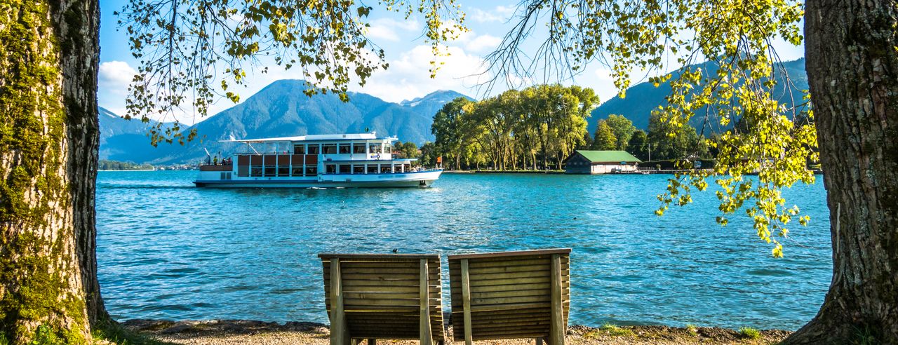 Zwei Liegestühle an einem See in Bayern, im Hintergrund sind Berge und auf dem See fährt ein Schiff