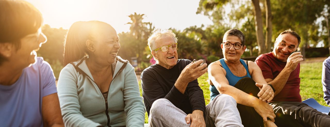 Eine Gruppe älterer Menschen sitzt auf Sportmatten in der Natur, trinkt Tee und sind glücklich während ihres Gesundheitsurlaubes
