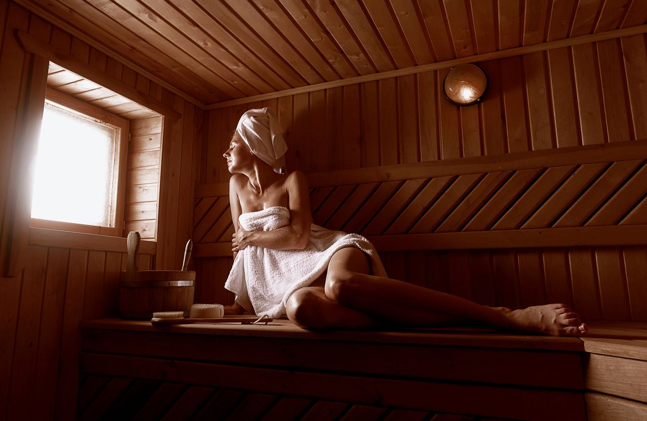 Eine Frau sitzt mit weißem Handtuch bekleidet in einer Sauna und hat ihren Blick durch ein kleines Fenster nach außen gerichtet. Vor ihr stehen ein Holzeimer und eine Holzbürste.