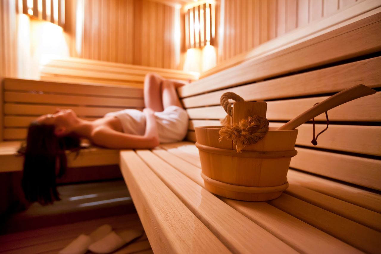 Eine Frau liegt auf dem Rücken in der Sauna und genießt Ihren Urlaub mit Sauna in NRW. Im Vordergrund steht ein Holzeimer auf der hölzernen Saunabank.