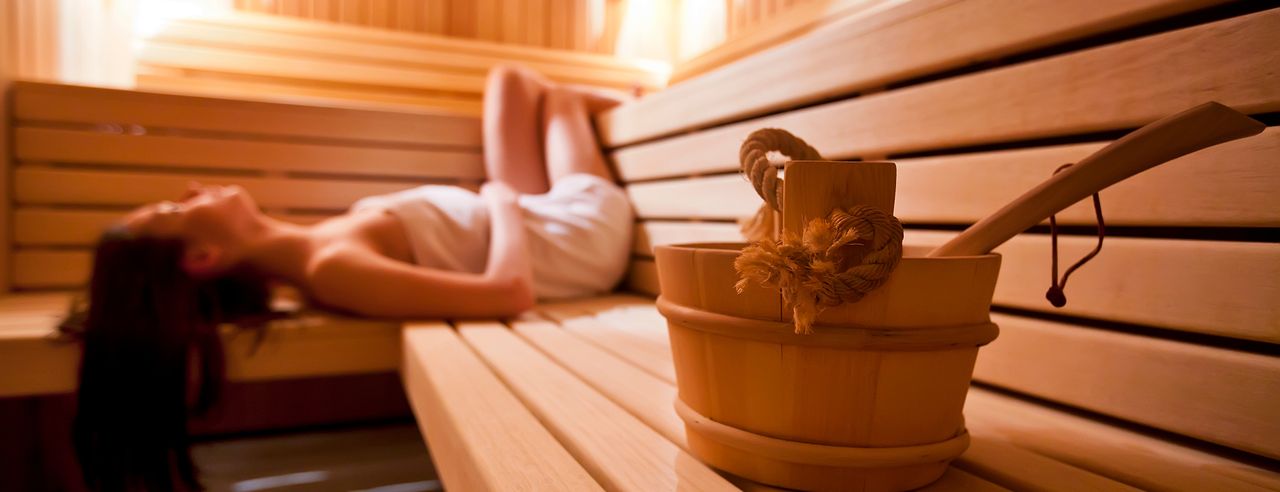 Eine Frau liegt auf dem Rücken in der Sauna und genießt Ihren Urlaub mit Sauna in NRW. Im Vordergrund steht ein Holzeimer auf der hölzernen Saunabank.