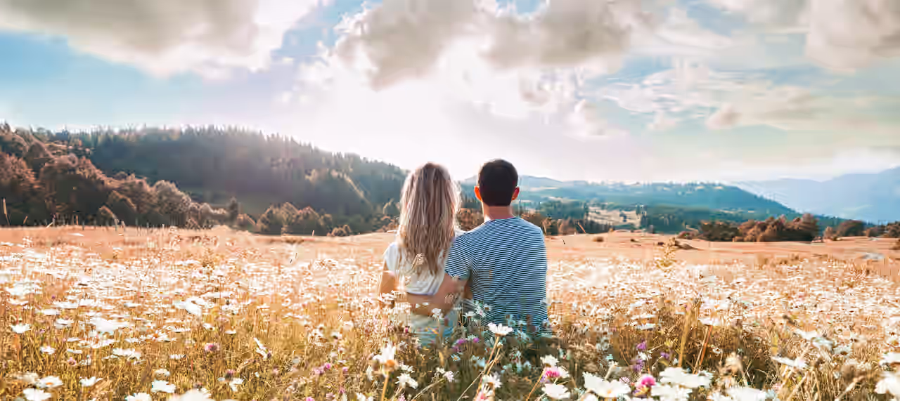 Ein Paar sitzt mit dem Rücken zu uns gedreht auf einer Blumenwiese und blickt auf die umliegende Natur.