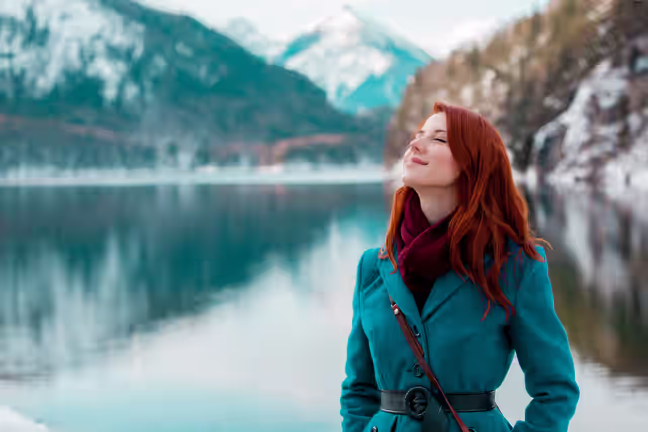 Eine Frau in türkisfarbener Jacke steht an einem See und hinter sind die Berge.
