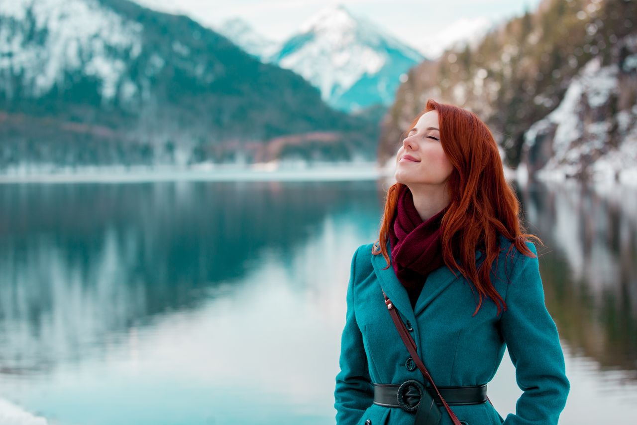 Eine Frau in türkisfarbener Jacke steht an einem See und hinter sind die Berge.