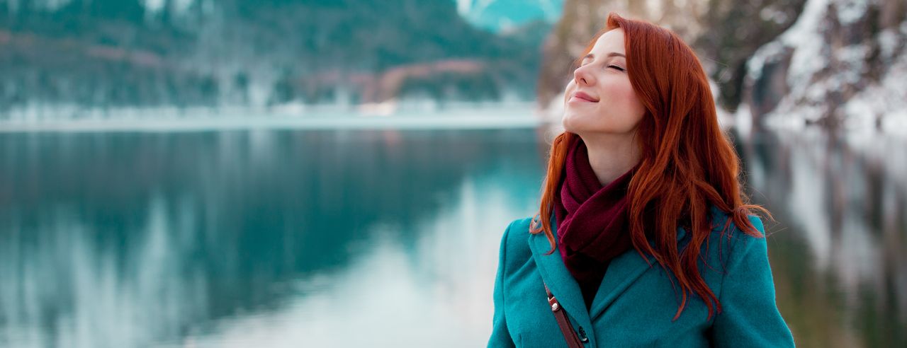 Eine Frau in türkisfarbener Jacke steht an einem See und hinter sind die Berge.