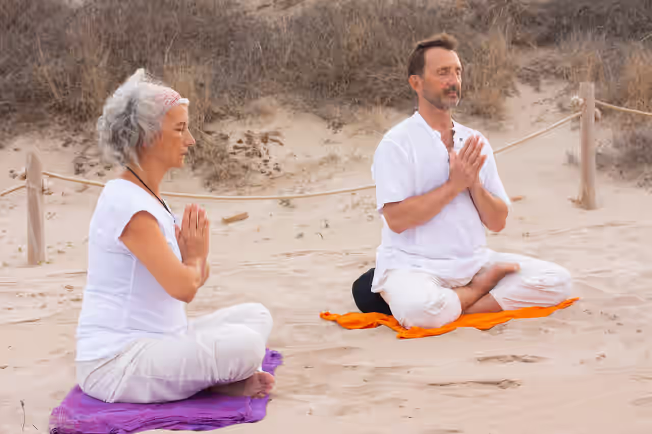 Zwei Personen mittleren Alters in weißer Kleidung praktizieren Kundalini Yoga auf bunten Handtüchern am Strand