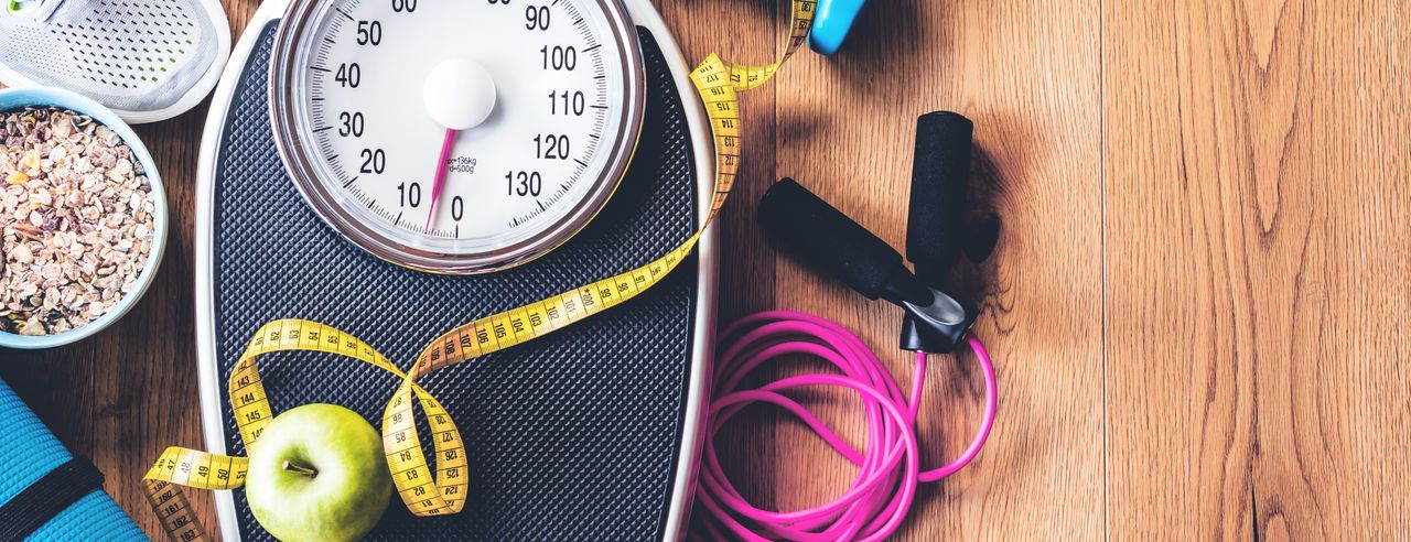 Scales with a yellow measuring tape on them, surrounded by fitness accessories such as dumbbells, skipping rope, running shoes and healthy food such as apples and oatmeal on a wooden floor.