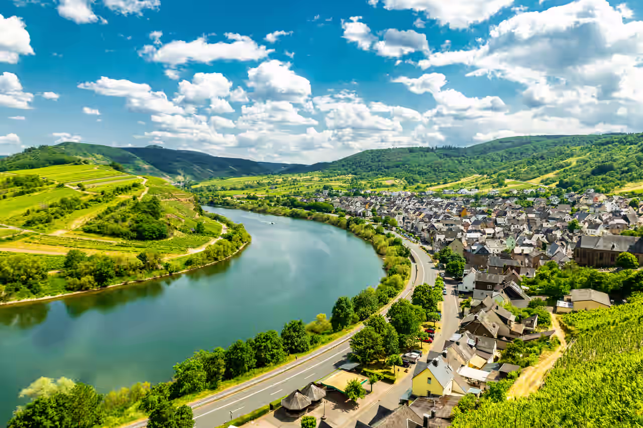 Blick auf die im Sonnenlicht glitzernde Mosel zwischen grünen Bäumen und Feldern und einer Ortschaft an der Straße