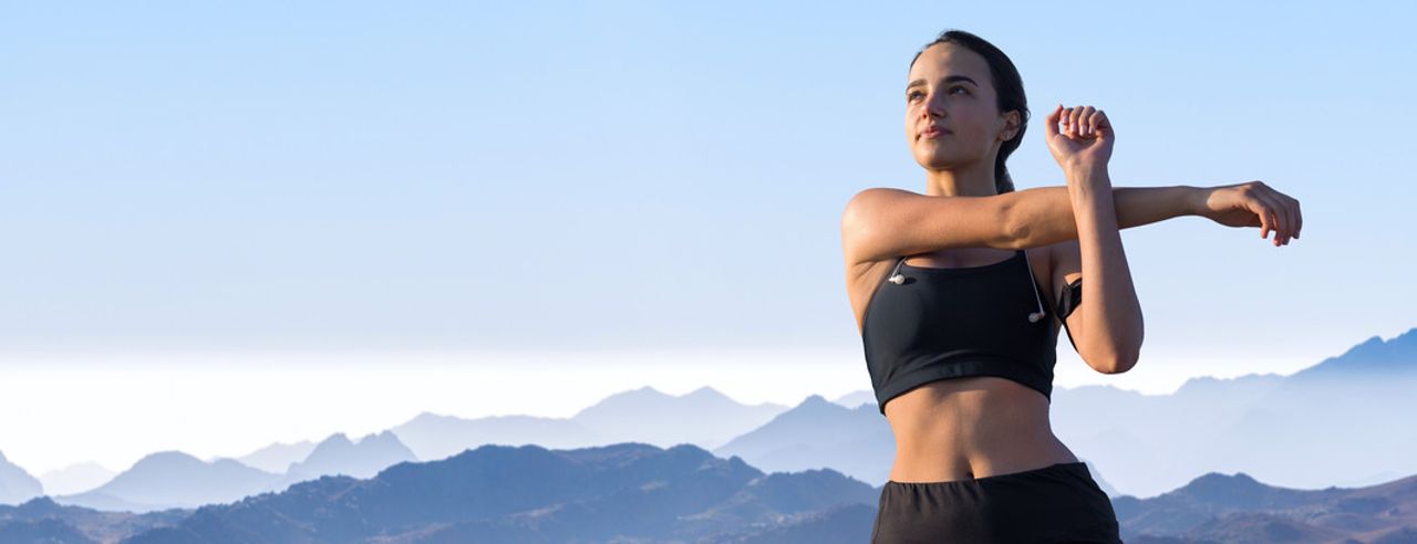 Frau in Sportkleidung dehnt sich, im Hintergrund Himmel und Berggipfel