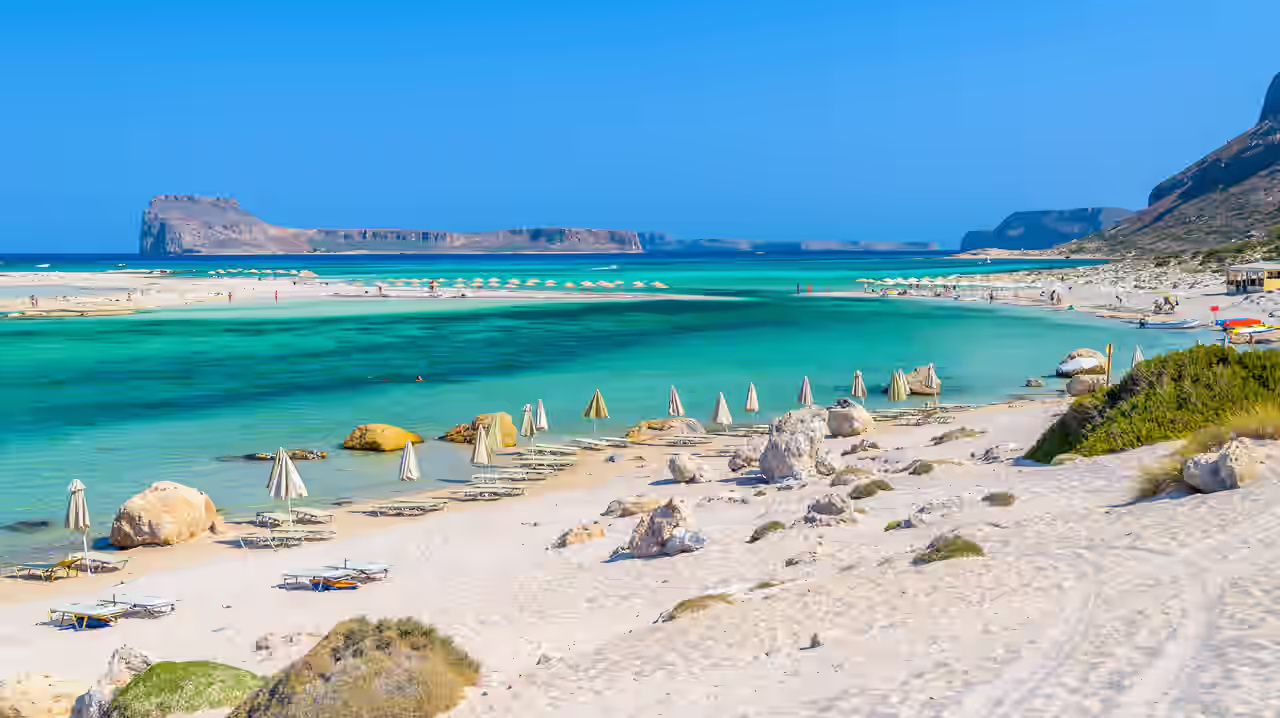 Sonnenschirme und Liegen am weißen Sandstrand mit großen Felsen an einer Bucht Kretas
