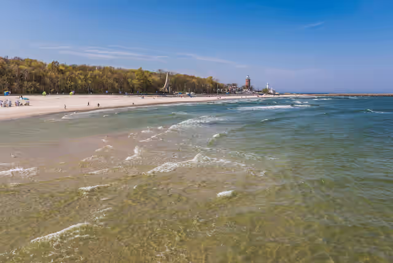 Blick über das klare Wasser der Ostsee auf den Strand mit Wald vor dem Leuchtturm von Kolberg