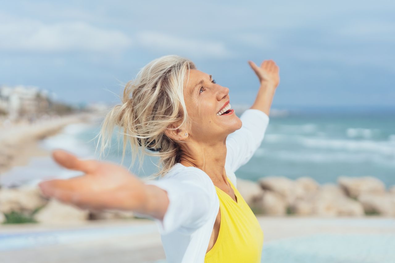Eine Frau in einem gelben T-Shirt steht am Meer und streckt freudig die Hände zu den Seiten aus.