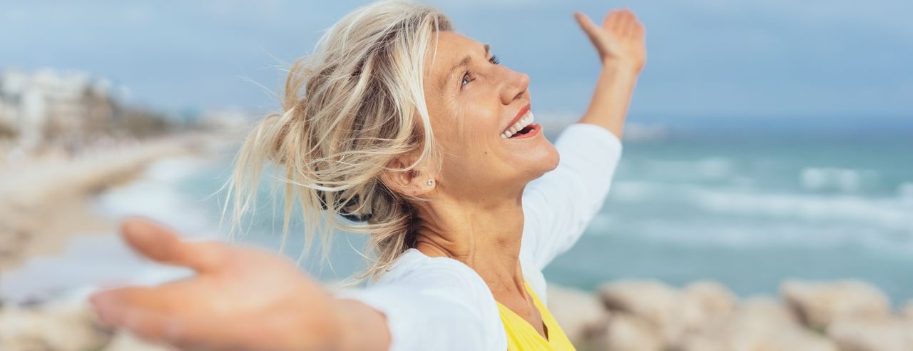 Eine Frau in einem gelben T-Shirt steht am Meer und streckt freudig die Hände zu den Seiten aus.