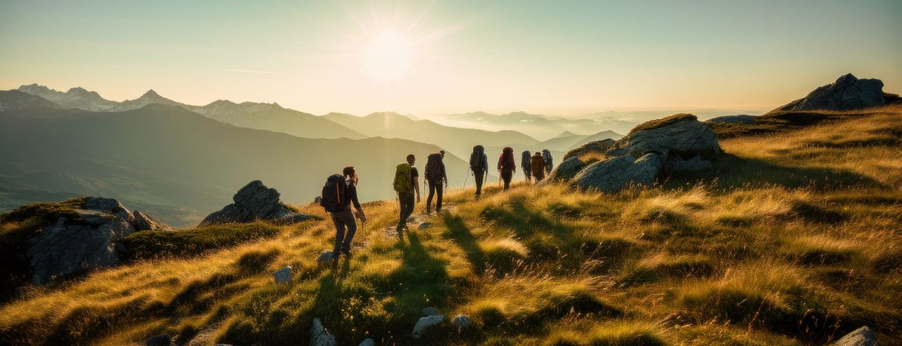 Eine Gruppe Wanderer wandert bei Sonnenuntergang auf einem mit Gräsern bewachsenen Berg, der von Sonnenlicht in warmes Gelb getaucht wird.