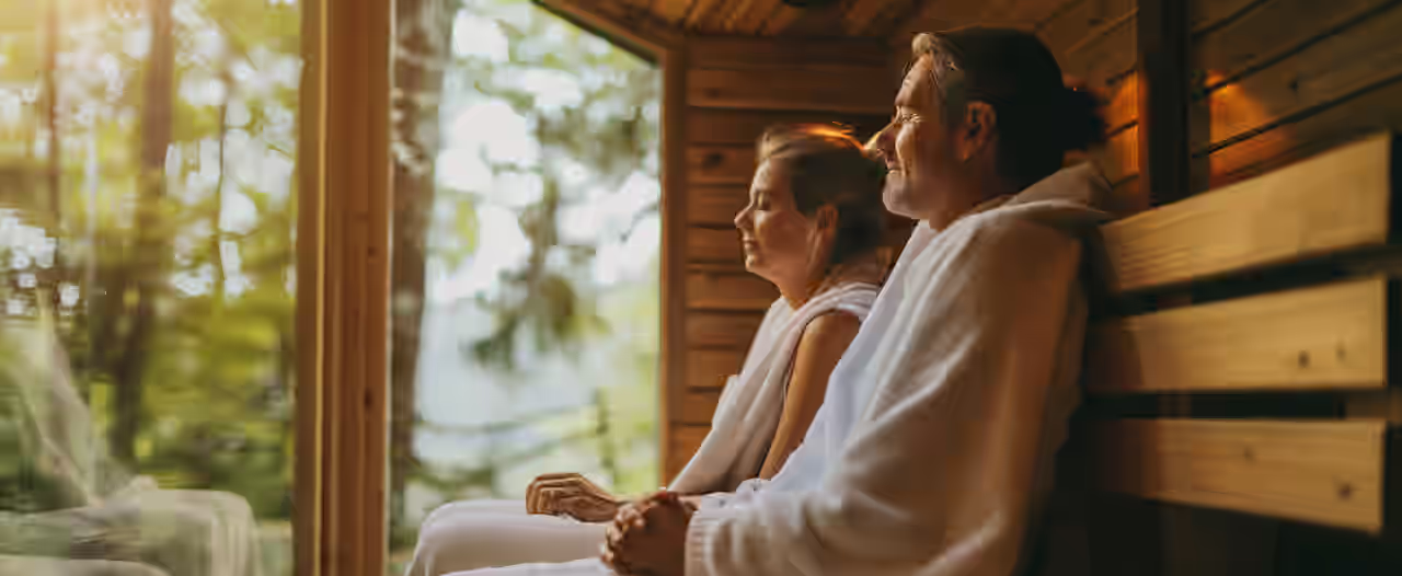 Ein Mann und eine Frau sitzen im weißen Bademantel mit geschlossenen Augen nebeneinander in einer mit Holz verkleideten Sauna und genießen die Nähe zur Natur, da die Sauna ein Panoramafenster hat und sich im Wald befindet