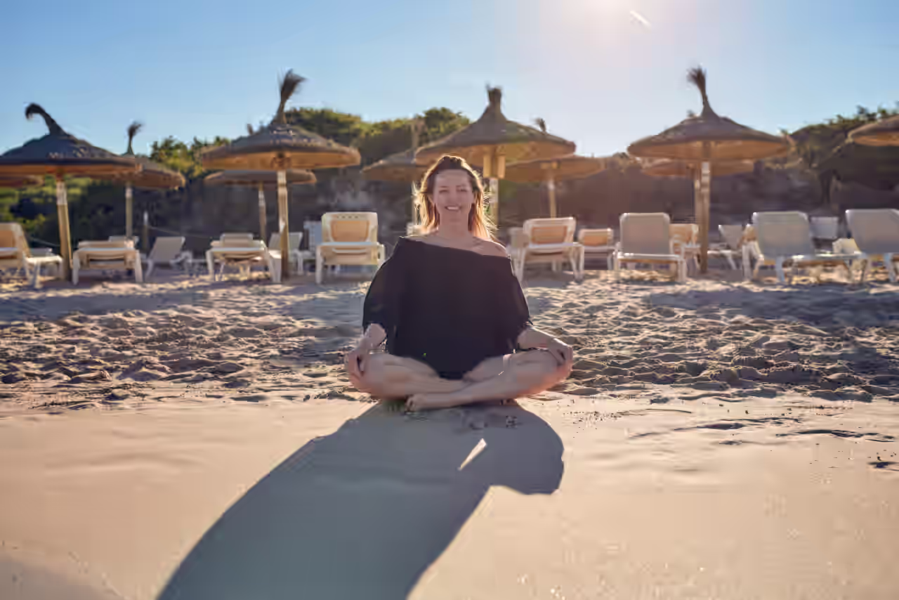 Frau sitzt im Schneidersitz am Strand vor Sonnenliegen und Sonnenschirmen