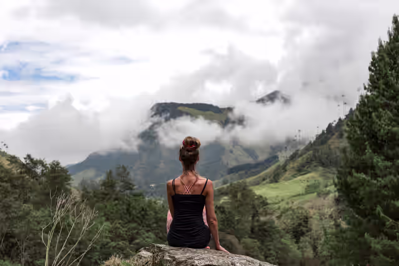 Frau sitz auf einem Berggipfel und guckt über von Wolken umrahmte Berge und Täler