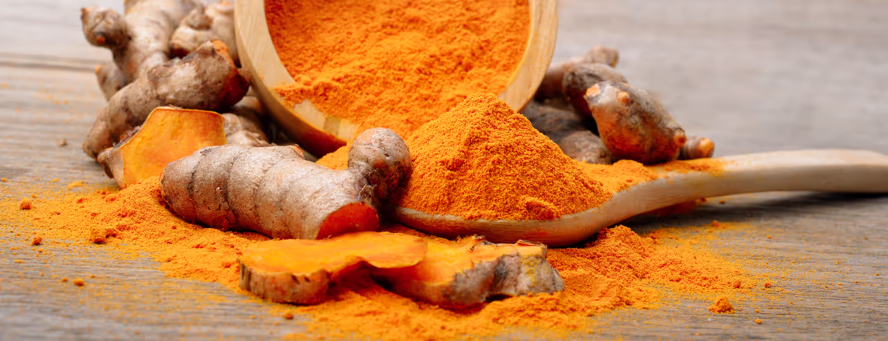 Su un tavolo di legno si trova una ciotola rovesciata contenente polvere di curcuma arancione. In primo piano un cucchiaio di legno e una radice di curcuma.