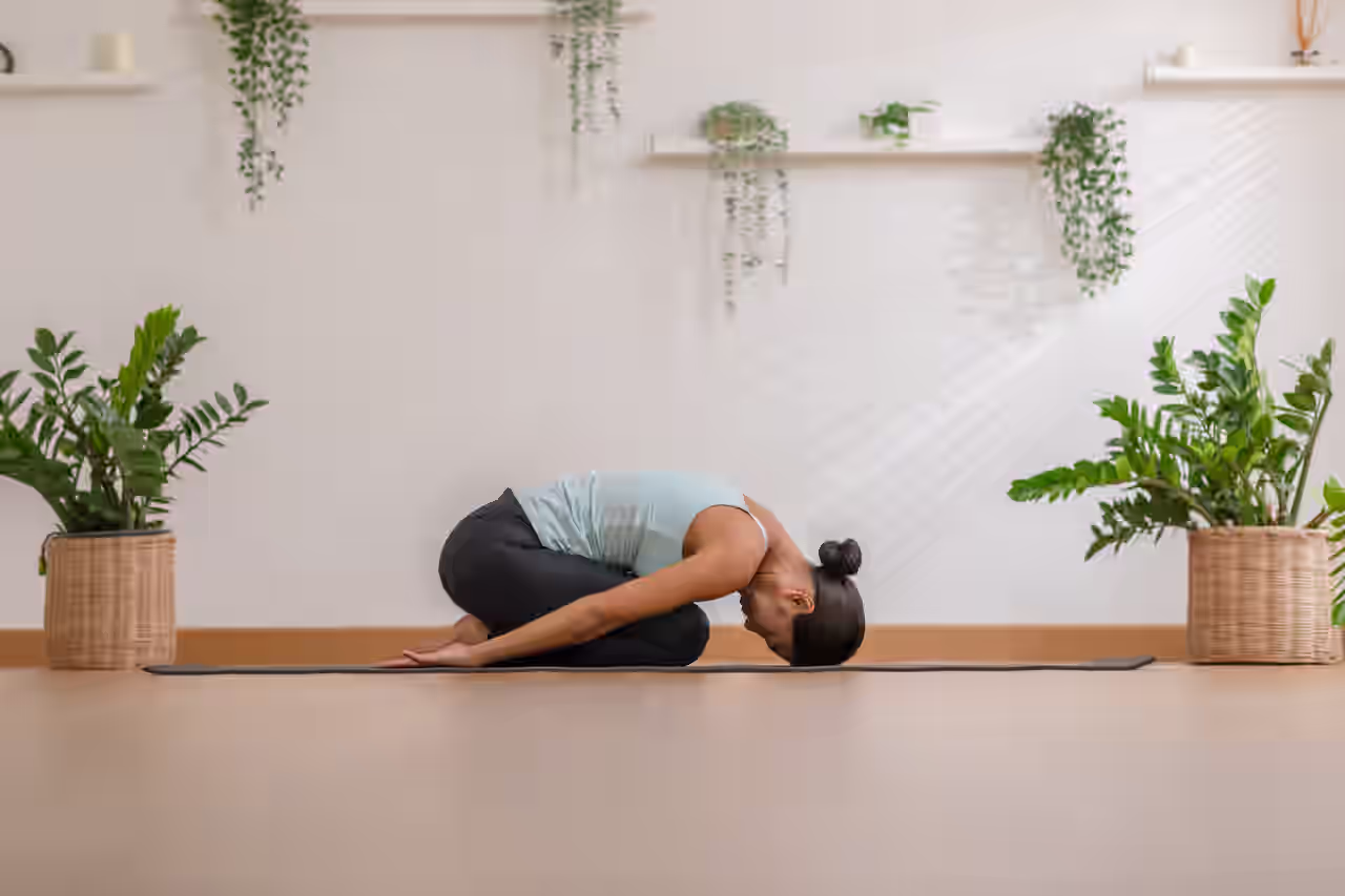Frau in der Haltung des Kindes auf einer Yogamatte zwischen geflochtenen Pflanzkübeln mit Grünpflanzen